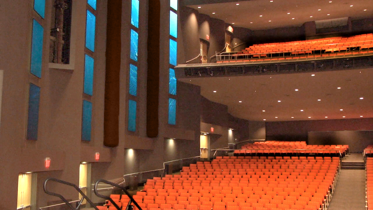 Modernes Theater mit orangefarbenen Sitzreihen, Balkonebene und hohen Wänden mit schmalen Fenstern.
