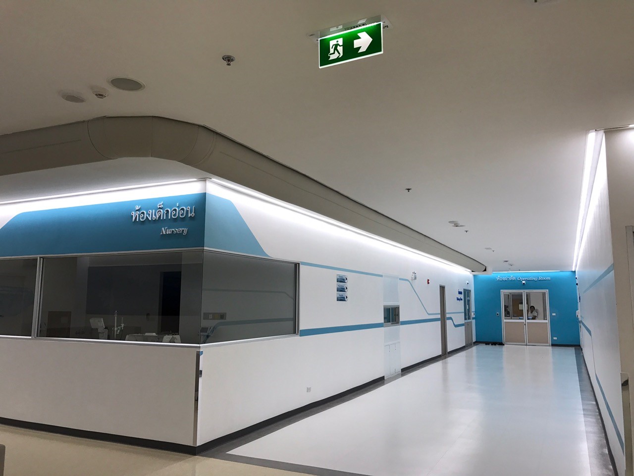 Krankenhausflur mit Empfangsbereich „Nursery“, klarer Linienführung und integriertem Textil-Luftkanal entlang der Decke.