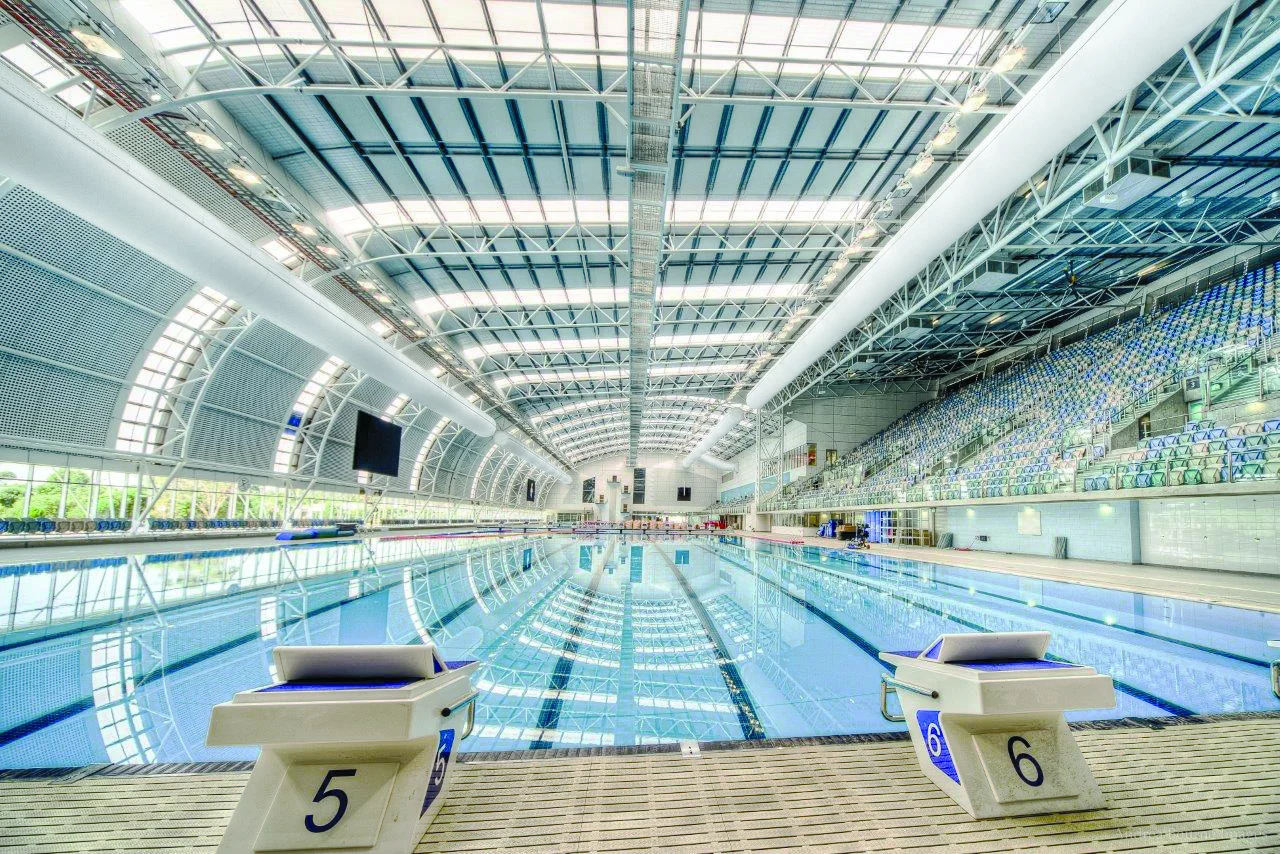 Große Hallenbadanlage mit abgehängten weißen Textilluftkanälen innerhalb der Stahl-Dachkonstruktion oberhalb des Schwimmbereichs.