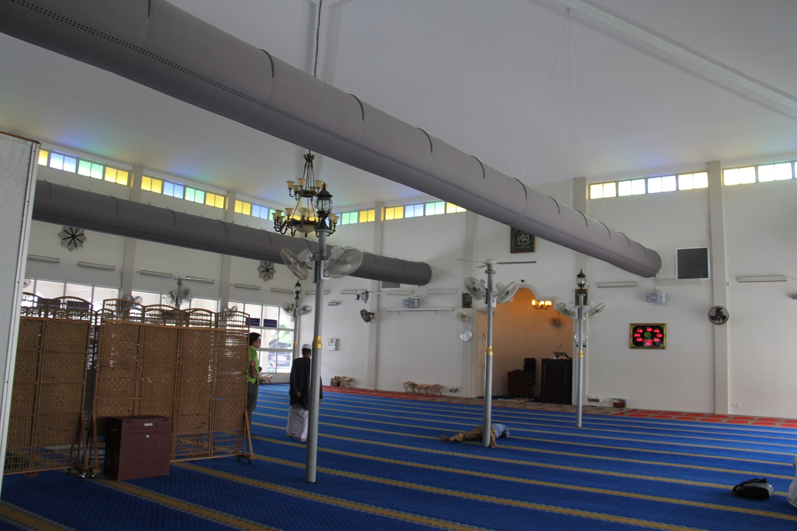 Gebetshalle mit langen grauen textilschlauch-Systemen unter der Decke, Säulenventilatoren und strukturiertem Teppichboden.