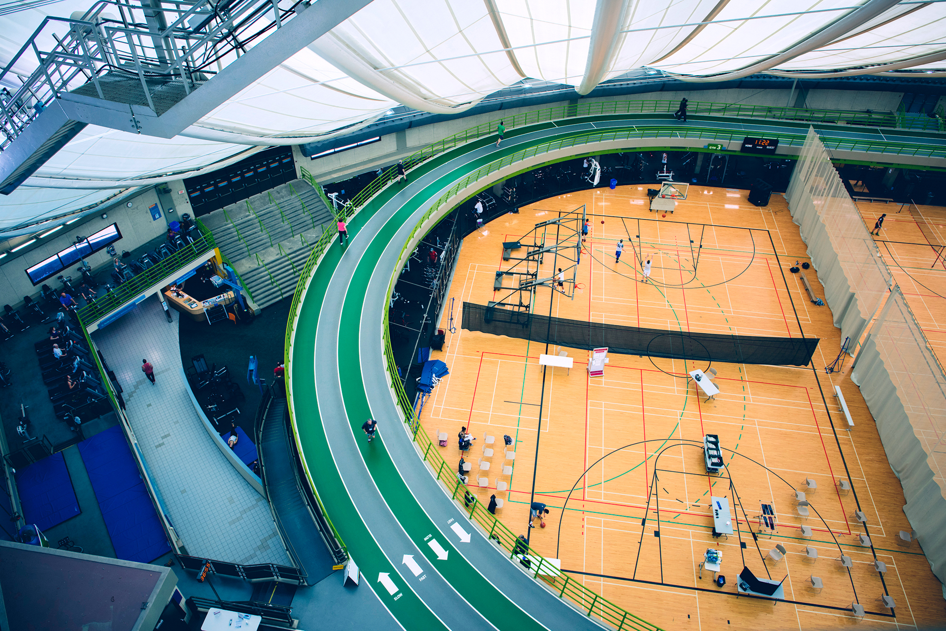 Moderner Indoor-Sportkomplex mit erhöhter Laufbahn und gespanntem textilen Deckensystem oberhalb der Basketballfelder.