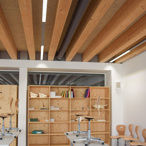 Klassenzimmer mit sichtbarem textilschlauch zwischen Holzträgern, kombiniert mit offenen Regalen und aufgestellten Stühlen nach Unterrichtsende.