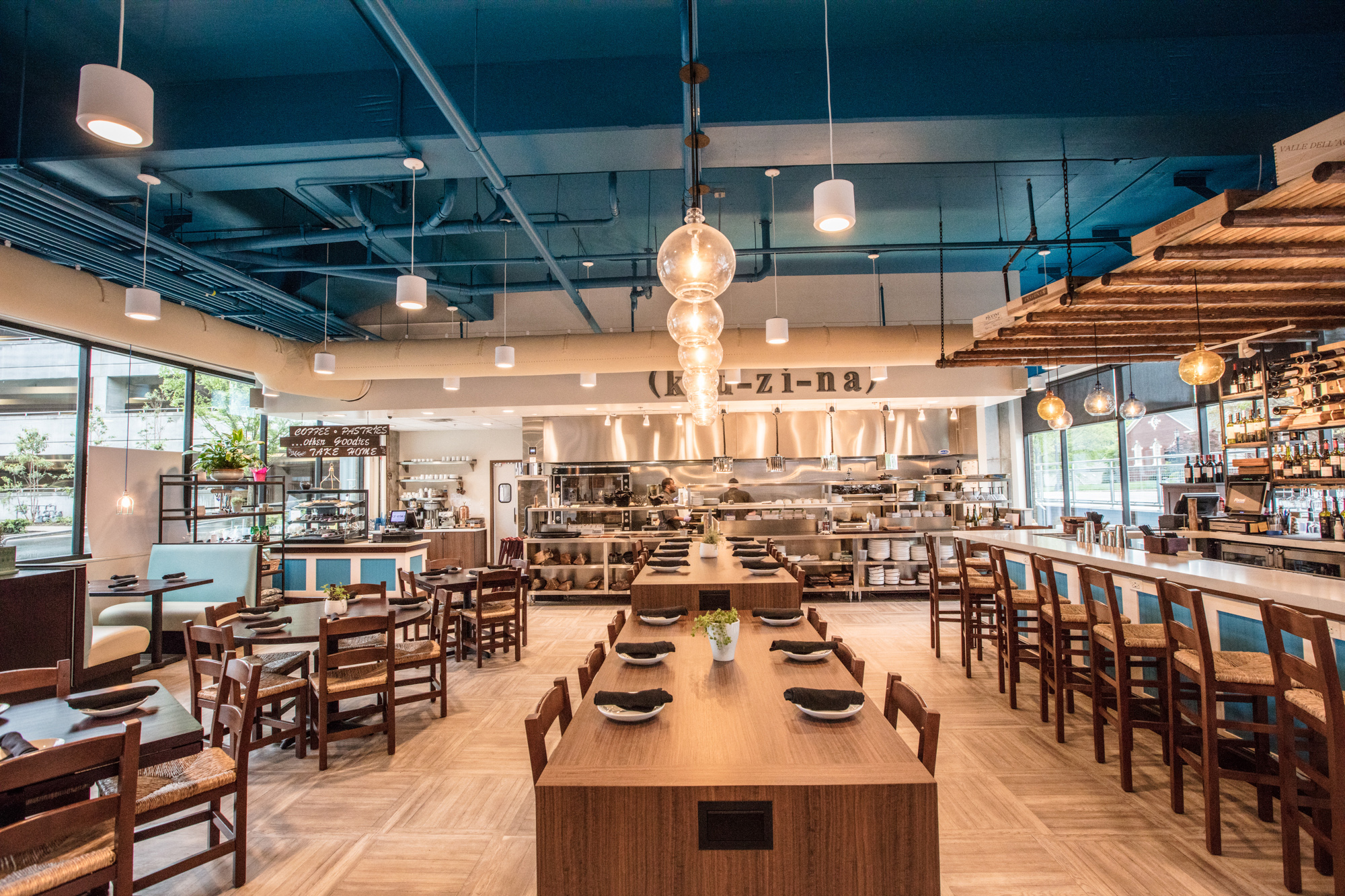 Modernes Restaurant mit sichtbaren textilen Luftkanälen, offener Küchenlandschaft und dekorativer Pendelbeleuchtung über langen Gemeinschaftstischen.