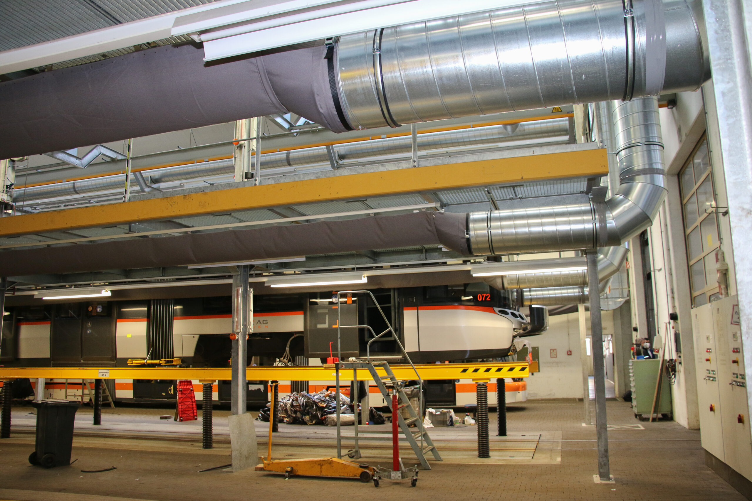 Industriehalle für Schienenwartung mit sichtbaren Textilluftkanälen und integrierten Metall-Lüftungssystemen oberhalb der Wartungsplattformen.