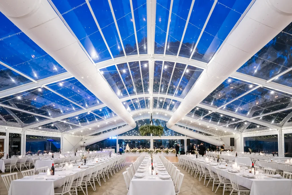 Salón de banquetes dentro de estructura de vidrio y metal, mesas largas blancas y decoraciones florales.