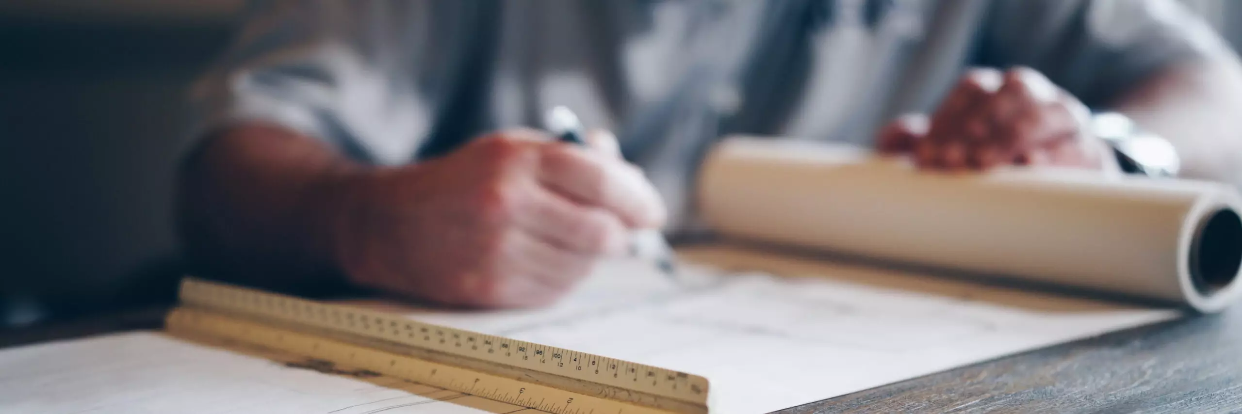 Person working on architectural drawings using a pen, triangular ruler, and rolled-up plans.